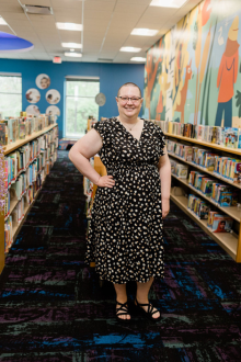 Jennifer (Jen) McArdle, Youth Services Librarian at Commerce Township Community Library, stands smiling in the children's library between rows of bookshelves. She is wearing a black dress with a white polka dot pattern and black sandals, with one hand on her hip. A colorful mural stretches along the wall behind her, creating a bright and welcoming library setting.